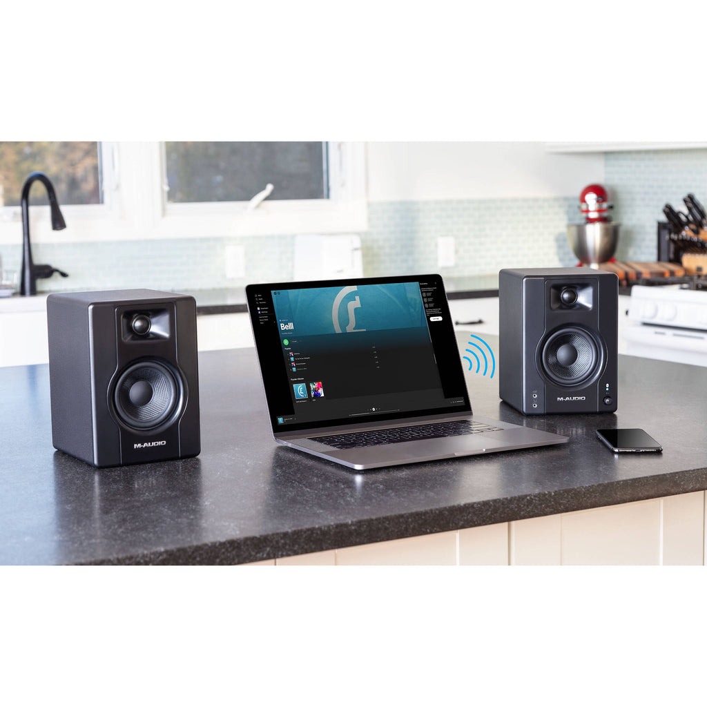 M-Audio BX3 BT desktop speakers showing black speakers with control knobs and connection ports on a desk surface.