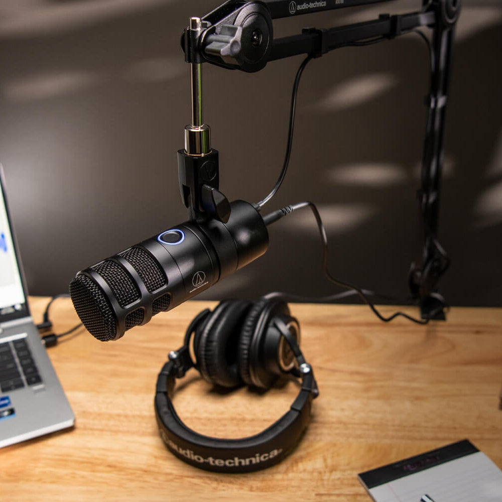Audio Technica AT2040USB dynamic USB microphone on a desk with a computer and headphones in a home recording setup.